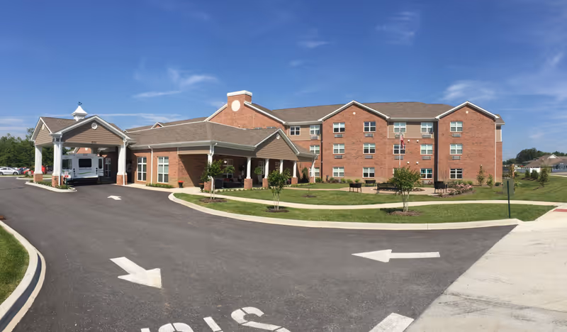 Front view of a three-story brick senior living building with a covered porte-cochère, driveway markings, and landscaped lawn under a blue sky.