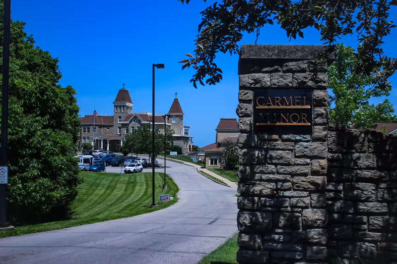 View of the entrance driveway to Carmel Manor, featuring a stone sign with the facility name and a large historic-style building with towers and crosses in the background under a clear blue sky.