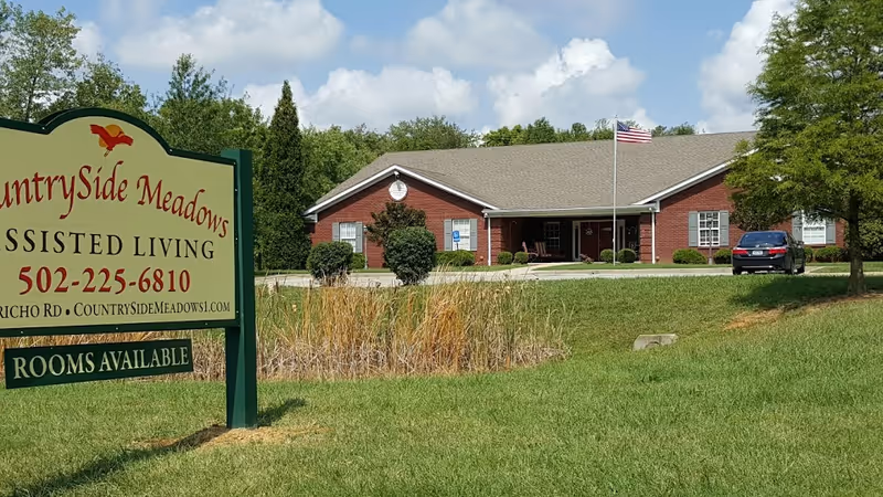 Exterior view of CountrySide Meadows assisted living facility showing a single-story brick building with a gray roof, an American flag on a flagpole, a parked car, and a green lawn with trees and shrubs. A sign in the foreground displays the facility's name, phone number, website, and availability of rooms.