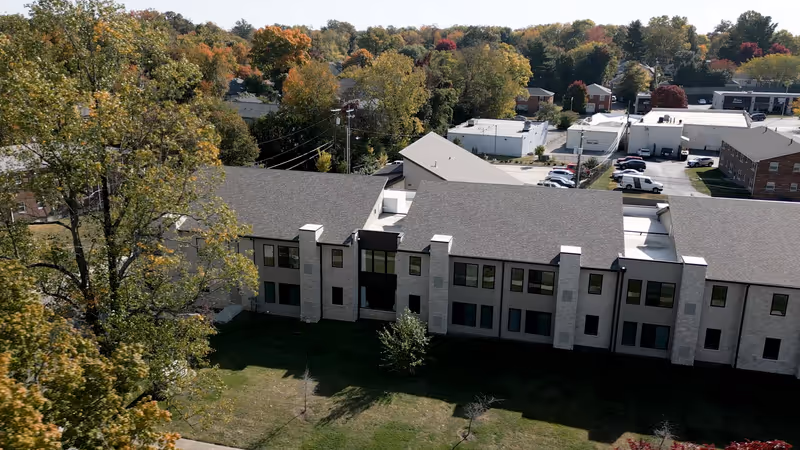 Aerial view of a long gray-brick senior living building with trees and a parking lot.