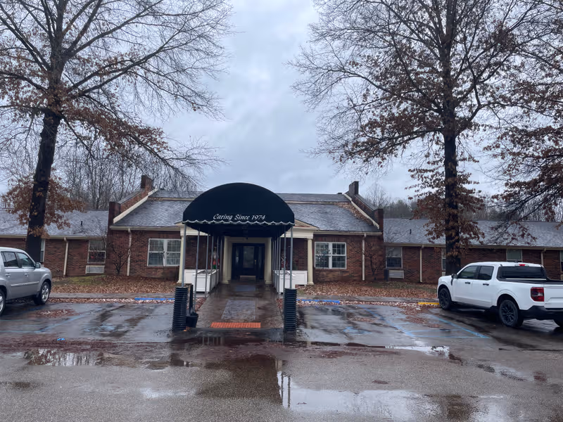 Brick senior living facility entrance with a curved canopy, wet pavement, and parked cars.