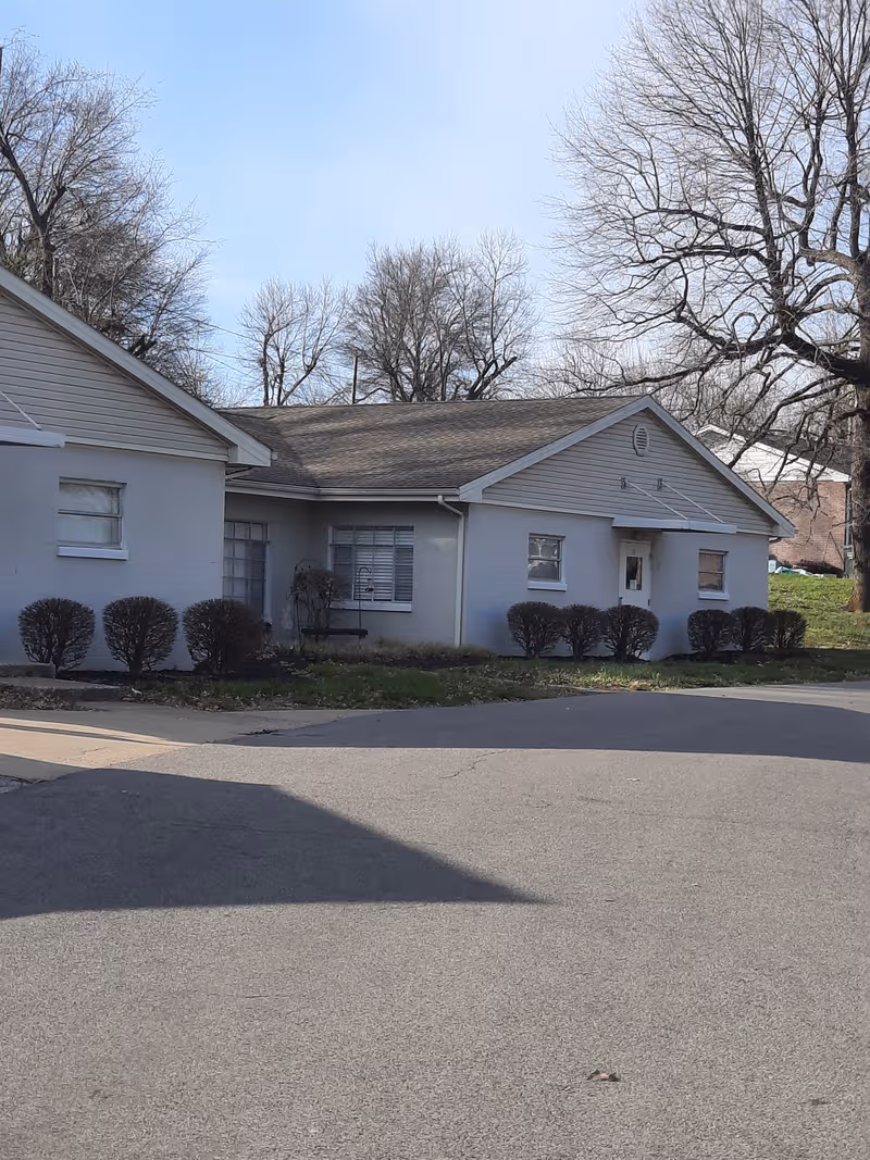 A single-story light-gray facility front with shrubs, leafless trees, and a paved driveway.