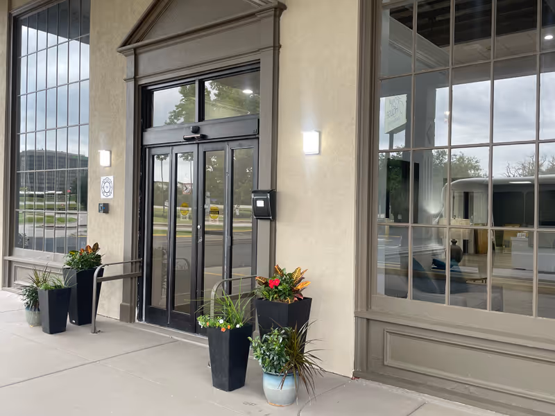 Entrance to The Spring House at Louisville with glass double doors framed by large windows. Several potted plants are placed on the concrete sidewalk near the entrance. The building exterior is beige with taupe trim, and there is a light fixture above the door.