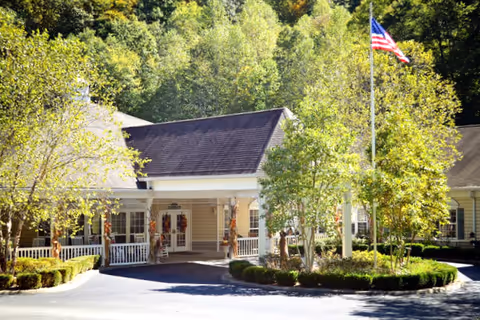 Exterior view of Cedar Creek Assisted Living facility showing a single-story building with a covered entrance, surrounded by trees and greenery. An American flag is flying on a flagpole near the entrance.