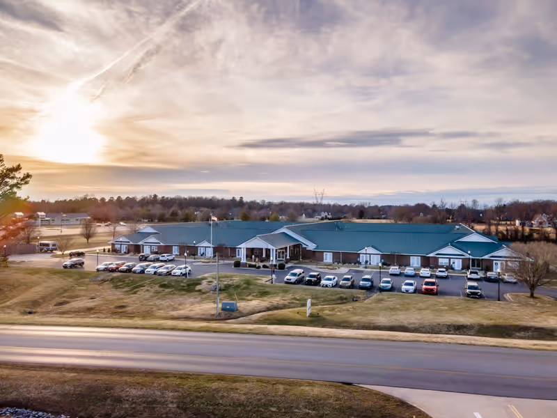 Aerial view of Rivercrest Place, a single-story senior living facility with a green roof and brick exterior, surrounded by a parking lot with several cars and open grassy areas under a partly cloudy sky during sunset.
