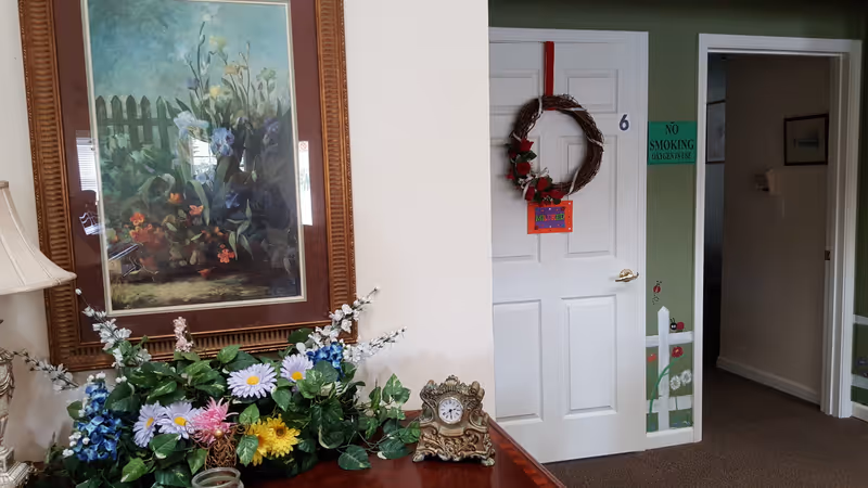 Interior view of a hallway in a senior living facility. A white door with the number 6 has a decorative wreath and a colorful sign that says 'Welcome'. To the left, there is a wooden table with a floral arrangement and an ornate clock. Above the table hangs a framed painting of flowers and a picket fence. The walls are painted green with a white chair rail and floral decorations near the base. A green sign on the wall reads 'No Smoking Oxygen in Use'.