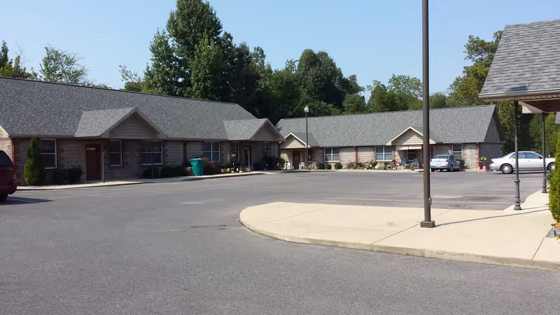 Exterior view of a single-story senior living facility with multiple connected units, a parking lot with several cars, and trees in the background under a clear blue sky.