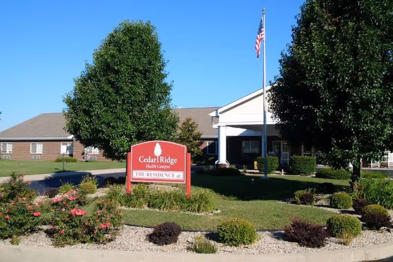 Front entrance of Cedar Ridge Health Campus showing a red sign, landscaped flowerbeds, a flagpole, and the building facade.