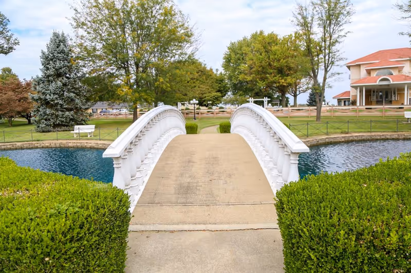 A white arched pedestrian bridge over a pond in a landscaped garden with trees and a building in the background.