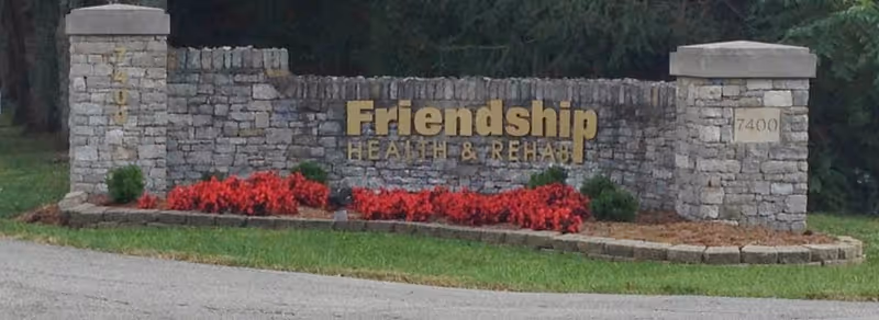 Stone entrance sign for Friendship Health & Rehab with red flowers planted in front and green grass surrounding it.