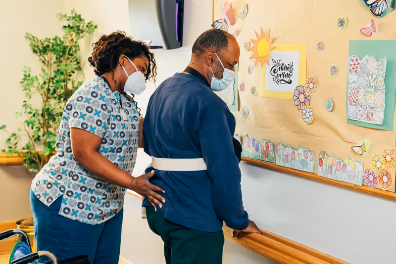 A caregiver wearing a patterned scrub top and a face mask assists an elderly man with a gait belt as he stands and looks at a bulletin board decorated with colorful spring-themed drawings and cutouts.