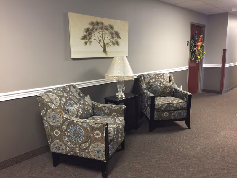 A hallway seating area in a retirement community featuring two patterned armchairs with matching cushions, a small black side table with a decorative lamp, and a wall painting of a tree. The hallway has beige and brown walls with white trim and a door decorated with a colorful floral wreath.