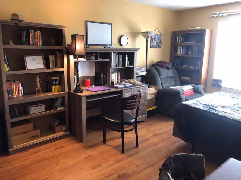 Cozy bedroom with wooden bookshelves and a desk, a recliner by the window, and a bed on wood flooring.