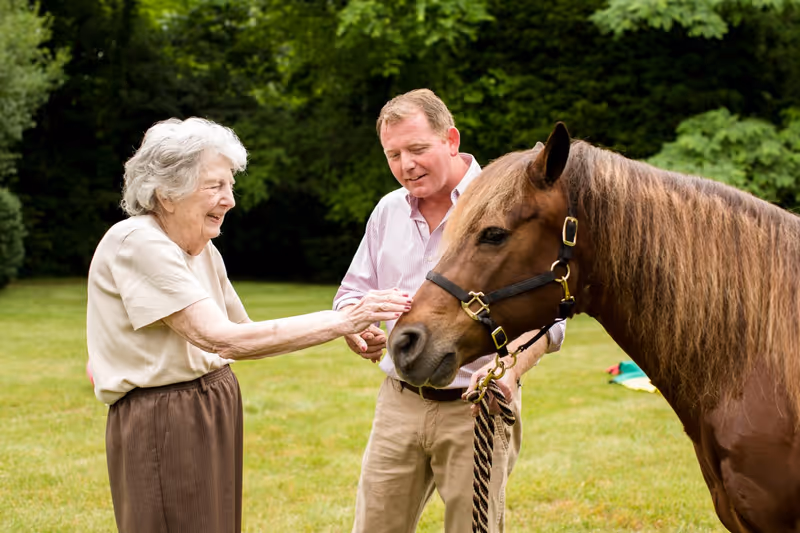 An elderly woman and a middle-aged man outdoors in a grassy area with trees in the background. The woman is smiling and reaching out to pet a brown horse with a black halter, while the man holds the horse's lead rope and looks on.