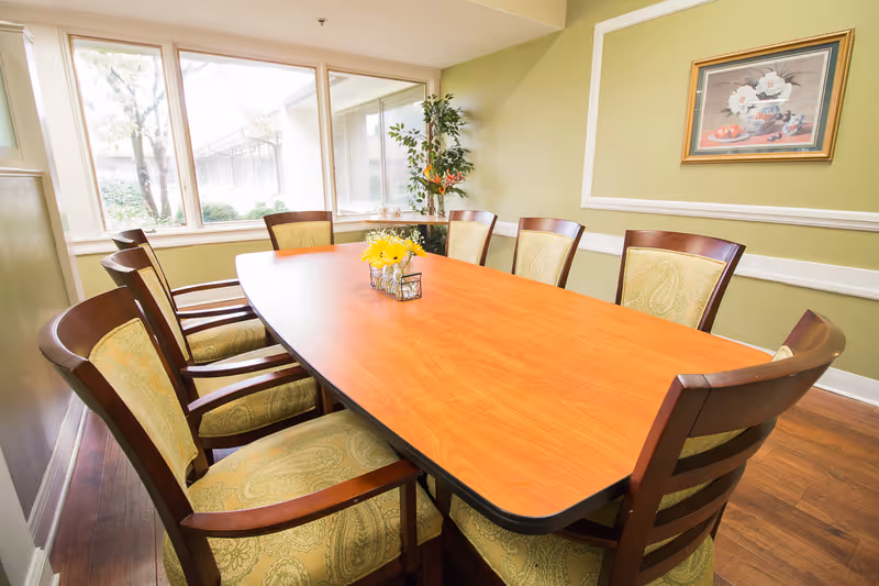 A dining room with a long wooden table surrounded by eight cushioned chairs with wooden frames. The walls are painted light green with white trim, and there is a framed floral painting on the wall. A small vase with yellow flowers is placed in the center of the table. Large windows let in natural light and show some greenery outside.