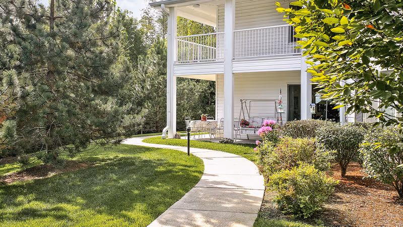 A curved concrete pathway winds through a well-maintained garden with green grass, bushes, and trees leading to a white two-story building with a covered porch. The porch has outdoor furniture including a swing and a table with chairs, surrounded by flowering plants and shrubs.