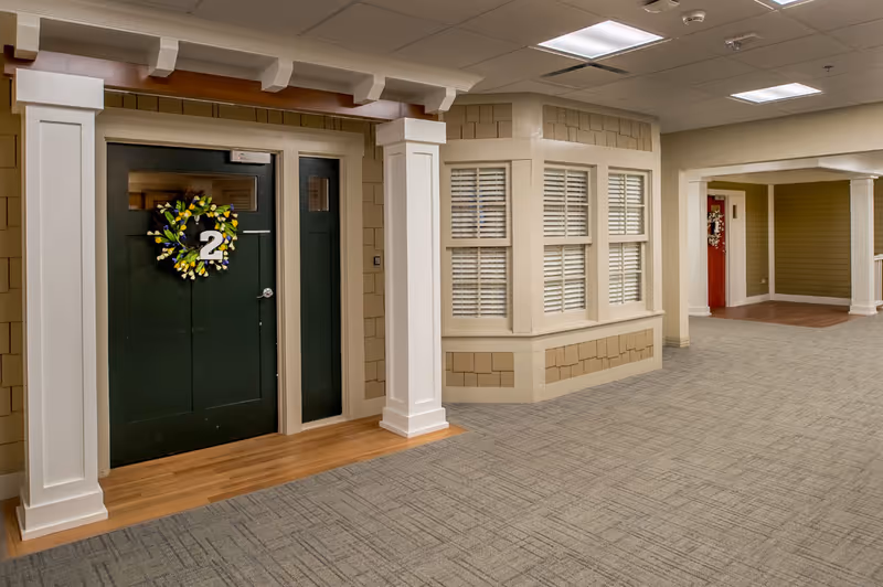 Interior hallway of a senior living facility with a green door marked with the number 2 and decorated with a floral wreath. The hallway features beige walls with white trim, a carpeted floor, and a window with white shutters. Another door with a red color and a wreath is visible further down the hallway.