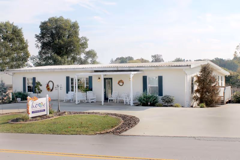 Front view of a single-story white assisted living building with a covered porch, chairs, landscaping, and a sign by the driveway.