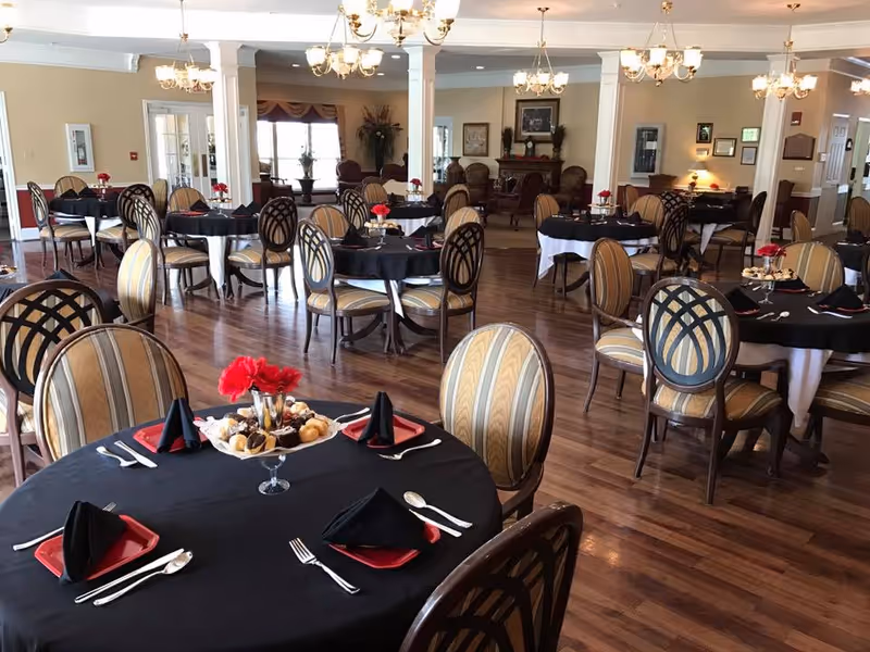A spacious dining hall with round tables, black tablecloths, striped chairs, and chandeliers.