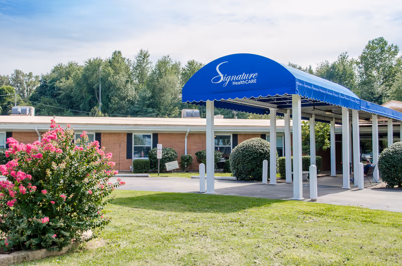 Exterior view of Signature HealthCARE at Hillcrest showing a single-story brick building with a blue awning entrance. The awning has the facility's name on it. There are bushes and flowering plants in the foreground and trees in the background under a partly cloudy sky.