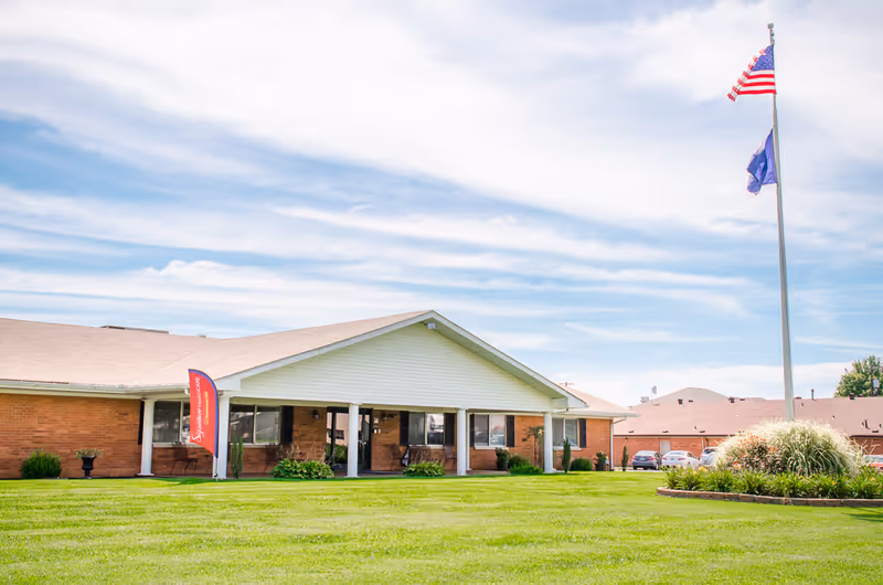 Exterior view of a single-story brick building with a white gabled roof and a covered porch area. There is a red vertical banner near the entrance that reads 'Signature HealthCARE'. A tall flagpole with the American flag and another flag stands on a well-maintained green lawn with a circular flower bed. Several parked cars are visible in the background under a partly cloudy sky.