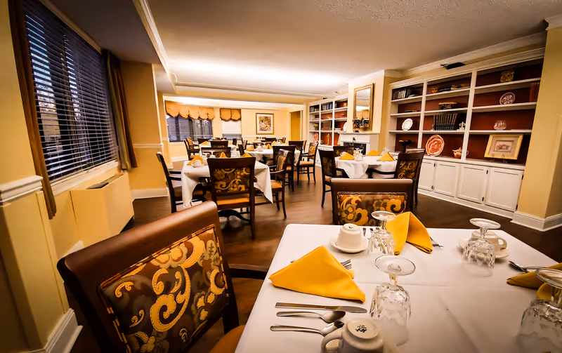 A warmly lit dining room with multiple tables covered in white tablecloths, each set with yellow folded napkins, upside-down glasses, cups, and silverware. The room features patterned chairs, large windows with blinds and curtains, and built-in shelves decorated with plates, books, and framed pictures.