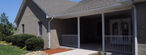 Exterior view of a single-story brick building with a covered porch supported by white columns, white railing, and a double door entrance. The building is surrounded by green bushes and a small landscaped area with mulch and rocks.