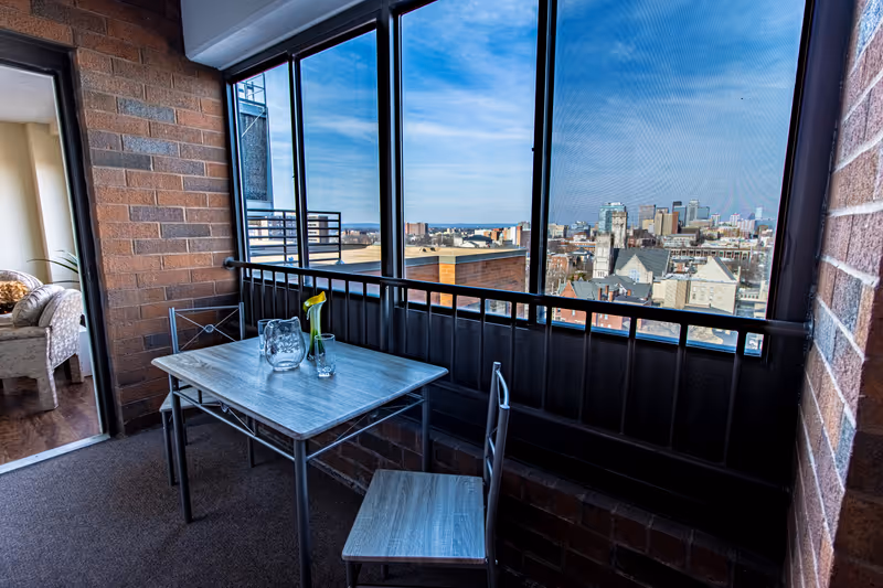 A small enclosed balcony area with a metal table and two chairs. On the table, there is a glass pitcher and a small vase with a single yellow flower. The balcony has large windows with a view of a city skyline under a blue sky. The interior of a living room with a beige armchair is visible through an open doorway.