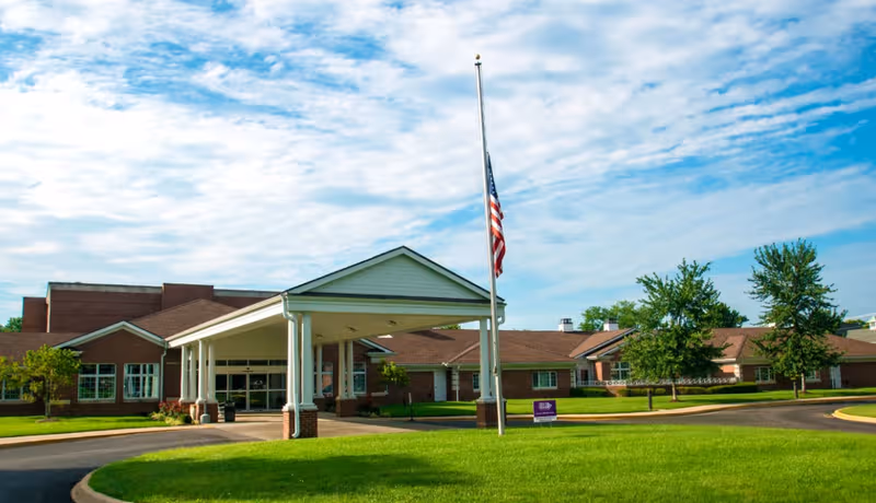 Exterior view of a senior living facility with a covered entrance, lawn, and flagpole.
