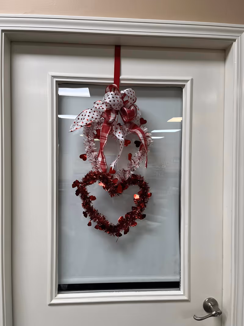 A white door with a glass panel in the center, decorated with two heart-shaped wreaths made of red and white tinsel and small red heart decorations. The wreaths are hung with a red and white polka dot and plaid ribbon tied in a bow at the top.
