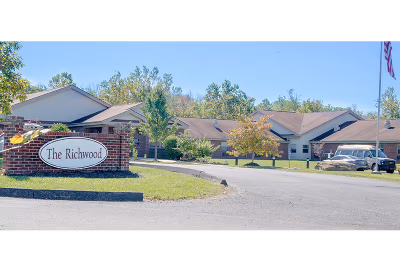 Front view of The Richwood nursing facility showing a brick entrance sign, single-story buildings, landscaping, and a parked vehicle.