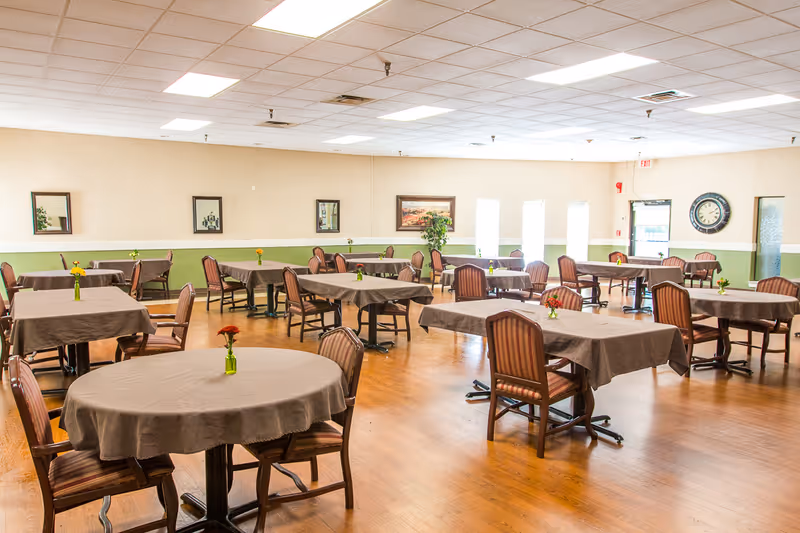 A spacious dining room with multiple tables covered in brown tablecloths, each table surrounded by wooden chairs with striped cushions. Small vases with flowers are placed on each table. The room has light-colored walls with a green accent stripe, framed pictures on the walls, a large clock, and bright overhead lighting. The floor is wooden.