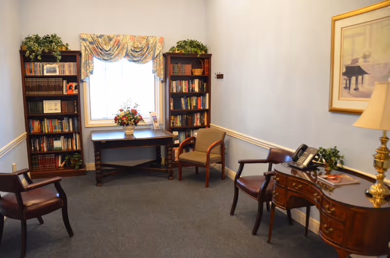 Library-style reading room in a senior facility, featuring tall bookcases, a window, plants, and seating.