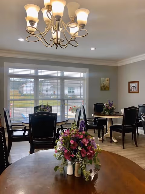 A well-lit dining room with round wooden tables and black chairs. Each table has a floral centerpiece with purple and pink flowers. Large windows with white blinds allow natural light to fill the room. A chandelier with multiple lights hangs from the ceiling. The walls are painted light gray and decorated with framed artwork.