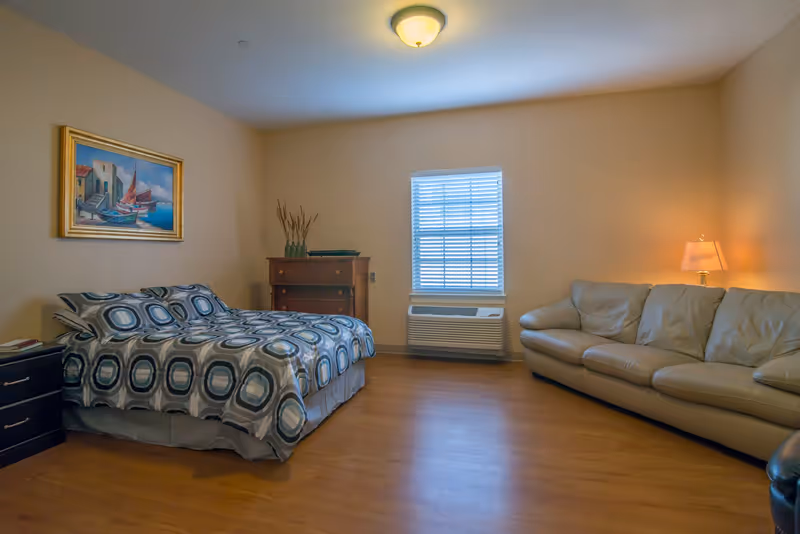 A bedroom with a bed covered in a patterned blue and gray bedspread, a wooden dresser with decorative vases, a window with blinds, a beige leather couch, a side table with a lamp, and a framed painting of boats on the wall.