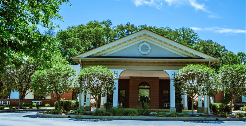 Front exterior view of Woodland Oaks Health Care building with a covered entrance, surrounded by neatly trimmed trees and greenery under a clear blue sky.