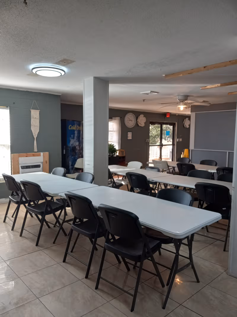 A bright dining room with long white tables and black folding chairs in a senior facility.