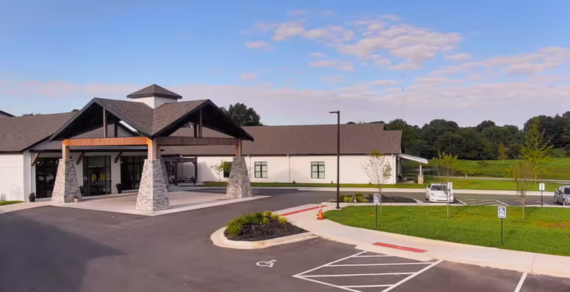 Front entrance of a single-story care facility with a covered portico, parking spaces, and landscaped lawn under a blue sky.