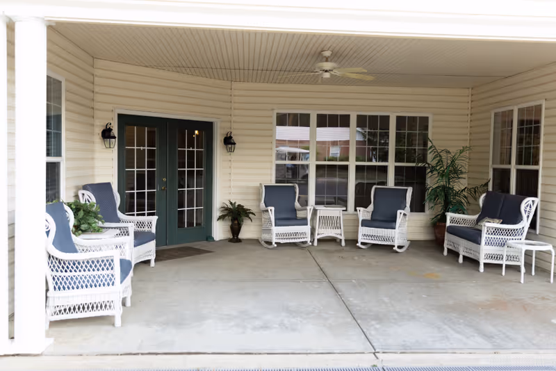 Covered entrance patio with white wicker chairs and blue cushions arranged around small tables in front of green double doors and windows.