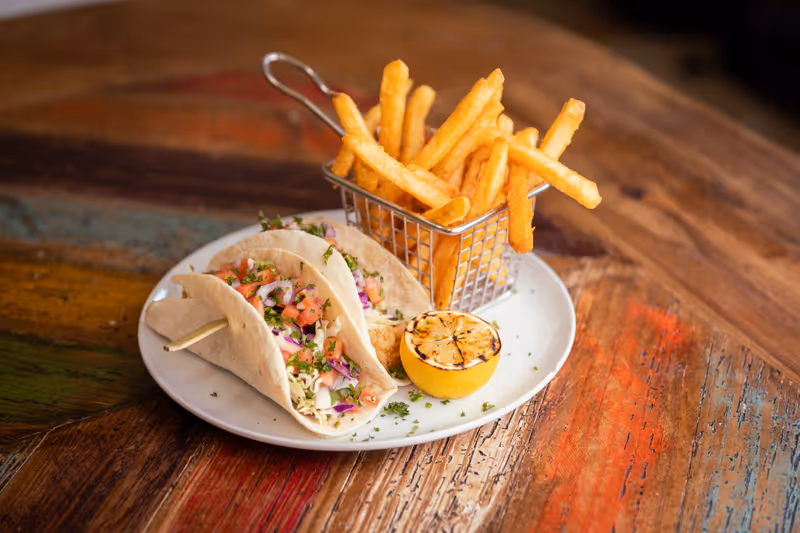 A white plate on a rustic wooden table with two soft tacos filled with chopped vegetables and herbs, a grilled lemon half, and a small metal basket filled with French fries.