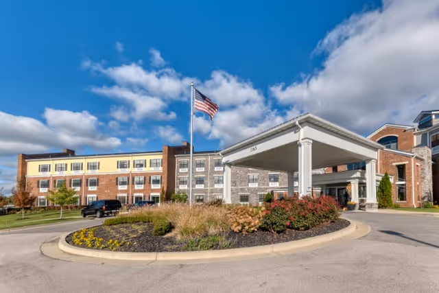 Exterior view of Cedarhurst of Beaumont, a multi-story senior living facility with a covered entrance driveway, landscaped roundabout with flowers and shrubs, and an American flag flying on a flagpole against a partly cloudy blue sky.