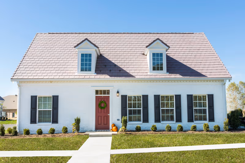 Front exterior view of a white brick house with a red door decorated with a green wreath, two pumpkins on the doorstep, black window shutters, and a gray shingled roof with two dormer windows under a clear blue sky.