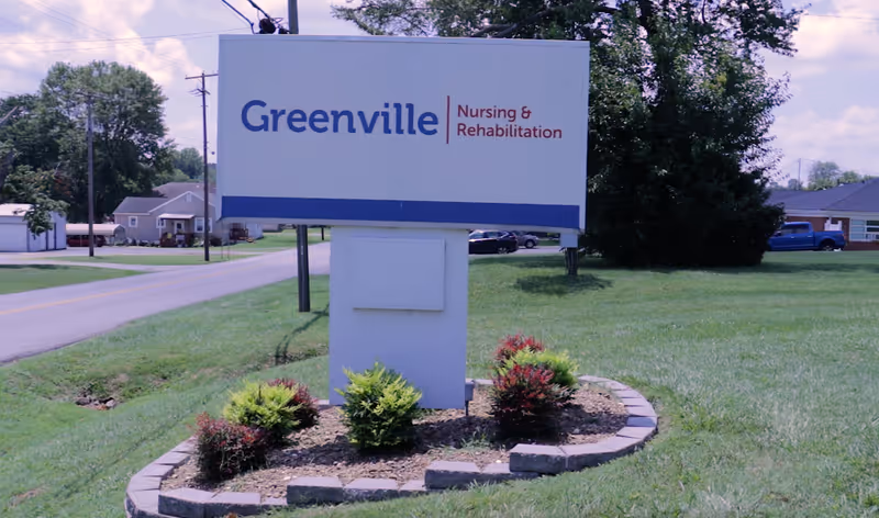 A freestanding roadside sign that reads "Greenville Nursing & Rehabilitation" on a grassy lawn with a road and houses in the background.