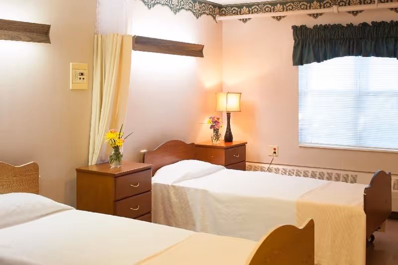 A bright bedroom with two single beds, wooden nightstands with flowers and a lamp, and a window with blinds.