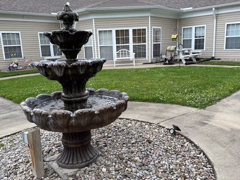 Outdoor courtyard area with a three-tiered stone water fountain surrounded by small rocks. In the background, there is a beige building with multiple windows and a white bench, a picnic table, and some outdoor chairs on a concrete patio. The area has green grass and a concrete walkway.