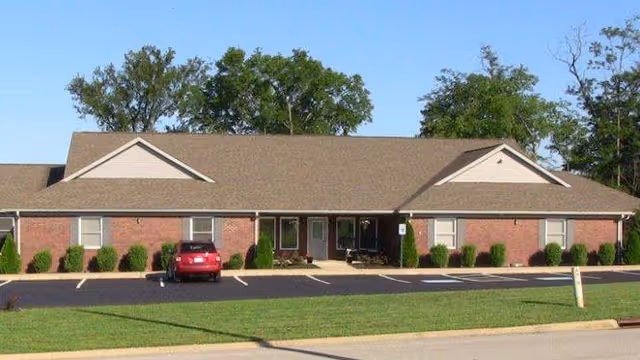 Single-story brick building with a brown shingled roof, several windows, and a small parking lot in front with one red car parked. The building is surrounded by green grass and some trees in the background under a clear blue sky.