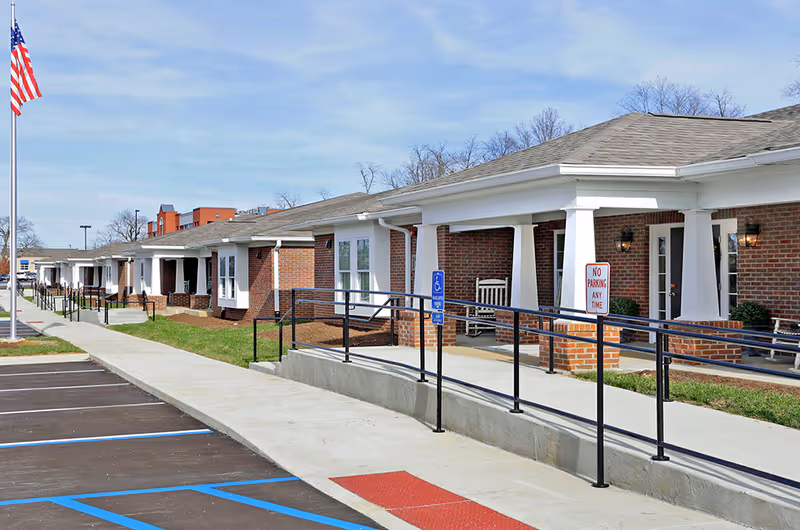 Single-story brick assisted living building with covered porches, a wheelchair ramp, parking spaces, and an American flag.