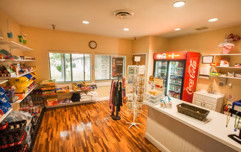 Interior view of a small convenience store area inside Florence Park Care Center featuring wooden flooring, shelves stocked with snacks and beverages, a Coca-Cola refrigerator, a counter with baked goods, and a rack with scarves and greeting cards near a window with blinds.