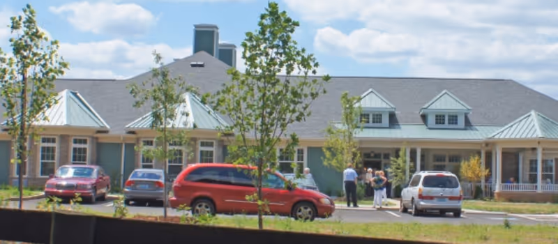 Exterior view of a single-story senior living facility building with a gray roof and light blue-green siding. Several cars are parked in front, and a few people are standing near the entrance. Young trees and landscaping are visible around the parking area under a partly cloudy sky.