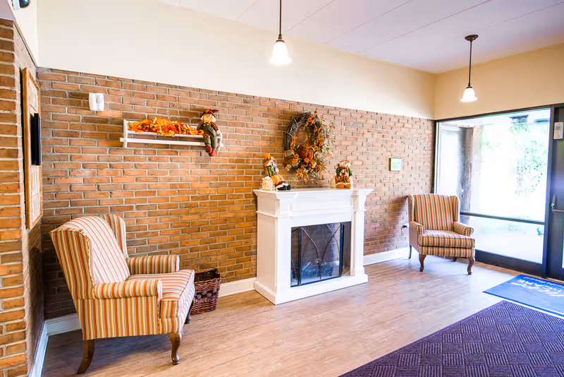 A cozy sitting area with two striped armchairs facing a white fireplace decorated with autumn-themed ornaments and a wreath on a brick wall. The room has wooden flooring, pendant lights hanging from the ceiling, and a glass door letting in natural light.